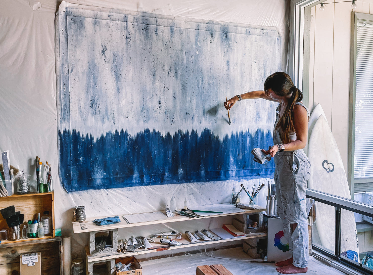 Artist and painter Liz C Fernandez painting a large 4 foot by 8 foot canvas.  The oil painting is abstract.  Dark blues rise to lighter blues with a white center line.  Liz C Fernandez abstract oil painter wears overalls in her painting studio. 