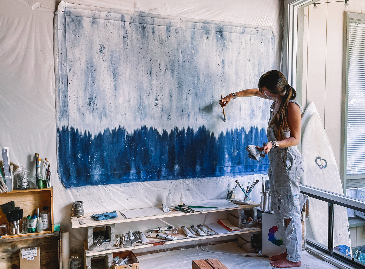 Artist and painter Liz C Fernandez painting a large 4 foot by 8 foot canvas.  The oil painting is abstract.  Dark blues rise to lighter blues with a white center line.  Liz C Fernandez abstract oil painter wears overalls in her painting studio.